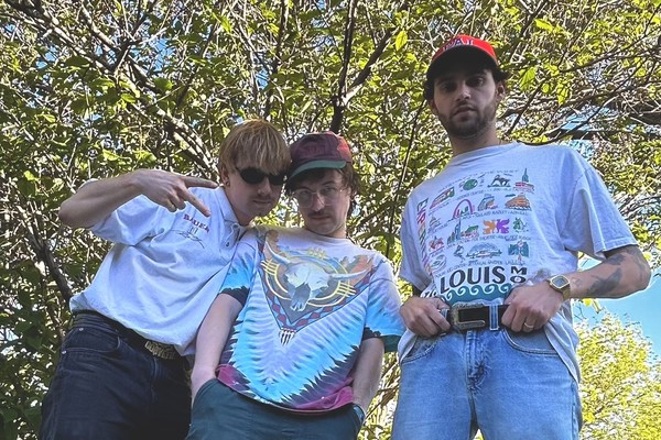 A colour photo shows three-piece band Tawni Bias posing in front of a leafy tree on a sunny day