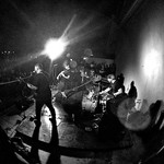 A black and white photo shows the band Taxa playing on stage under bright white lights