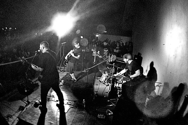 A black and white photo shows the band Taxa playing on stage under bright white lights