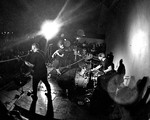 A black and white photo shows the band Taxa playing on stage under bright white lights