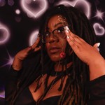 Hip-hop artist Tea Fannie poses with her hands touching her glasses in front of a bright wall with neon pink hearts