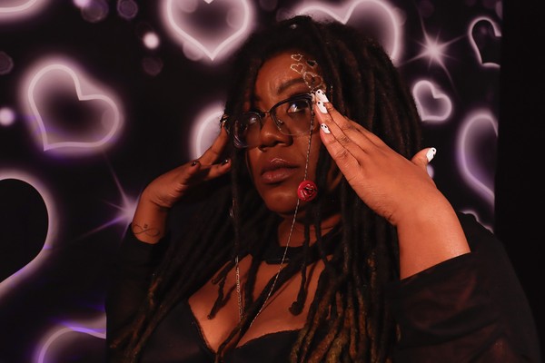 Hip-hop artist Tea Fannie poses with her hands touching her glasses in front of a bright wall with neon pink hearts