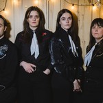 A colour photo of four-piece group The Garrys standing against a wooden planl wall with strings light strewn across. All member are wearing western type shirts with bolo tyes.