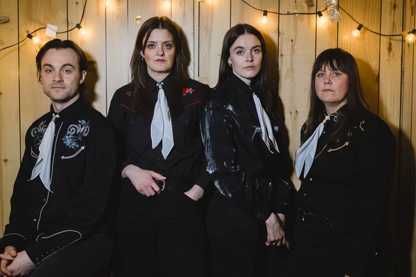 A colour photo of four-piece group The Garrys standing against a wooden planl wall with strings light strewn across. All member are wearing western type shirts with bolo tyes.