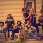 A colour photo shows seven-piece country band posing outside a house. All are in western wear.
