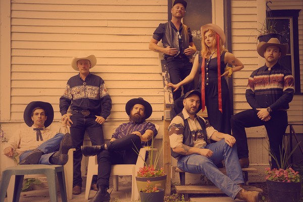 A colour photo shows seven-piece country band posing outside a house. All are in western wear.
