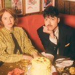 A colour photo shows two members of the group Thomas Thomas sitting inside a red leather booth with an uneaten cake sitting on the table in front of them