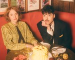 A colour photo shows two members of the group Thomas Thomas sitting inside a red leather booth with an uneaten cake sitting on the table in front of them