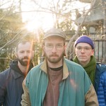 A colour photo shows the three members of UTILS standing outdoors in a lightly wooded area with leafless branches and softly lit by the setting sun in the background. The member in the foreground wears glass, a baseball cap, and a green and tan jacket. Behind them are the two other members, one wearing a dark jacket and the other wearing a blue puffer jacket, purple toque, and a green scarf.