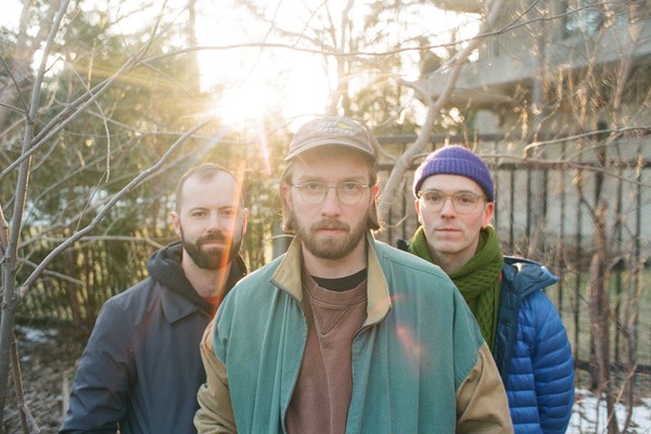 A colour photo shows the three members of UTILS standing outdoors in a lightly wooded area with leafless branches and softly lit by the setting sun in the background. The member in the foreground wears glass, a baseball cap, and a green and tan jacket. Behind them are the two other members, one wearing a dark jacket and the other wearing a blue puffer jacket, purple toque, and a green scarf.