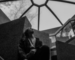 A black and white photo shows artist Void Comp sitting in a concrete room with a glass ceiling. 