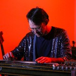 A colour photo shows musician Wayne Patrick Garrett playing a lapsteel with an orange background behind.