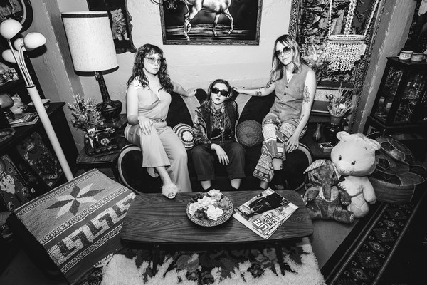 A black and white photo shows the three-piece band Window Lamp Corp posing on a couch