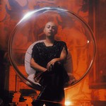 A colour photo of artist ZENON shows them sitting in a clear pod chair in an orange lit room.
