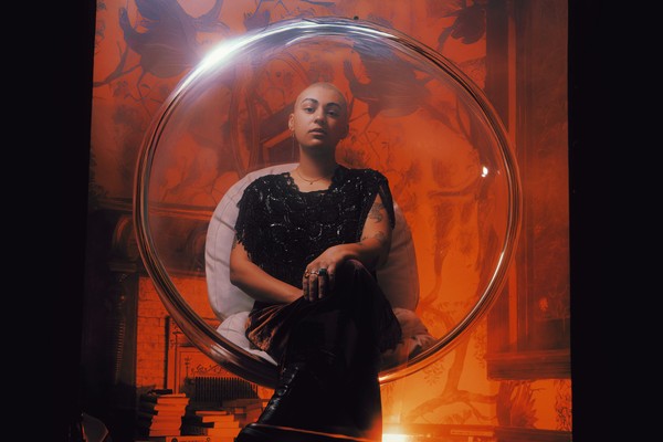 A colour photo of artist ZENON shows them sitting in a clear pod chair in an orange lit room.
