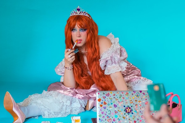 A colour photo of artist SHY FRiEND shows them sitting in a blue room with smudged makeup lighting a cigarette. Hands stick out at the forefront all handing out their bank cards.