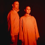The duo Bibi Club is seen standing under an orange light so they both appear completley orange. There is a dark black background behind them.