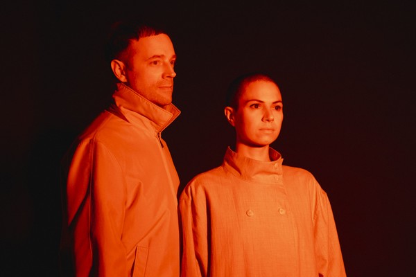 The duo Bibi Club is seen standing under an orange light so they both appear completley orange. There is a dark black background behind them.