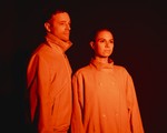 The duo Bibi Club is seen standing under an orange light so they both appear completley orange. There is a dark black background behind them.