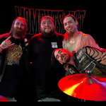 A colour photo shows four-piece band No More Moments posing for a photo under a red stage light. You can see a symbol from a drumkit in front of the group, and a black No More Moments banner behind them.