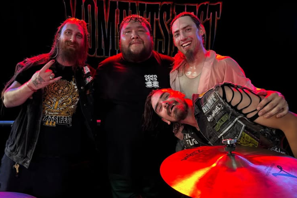 A colour photo shows four-piece band No More Moments posing for a photo under a red stage light. You can see a symbol from a drumkit in front of the group, and a black No More Moments banner behind them.