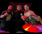 A colour photo shows four-piece band No More Moments posing for a photo under a red stage light. You can see a symbol from a drumkit in front of the group, and a black No More Moments banner behind them.