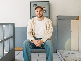 Portrait of comedian Nathan Hare seated in a lightly furnished room with a cube shelf beside him.