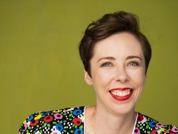 Portrait of comedian Luba Magnus wearing a vibrant patterned top,  smiling toward the camera, with a neutral background behind them.