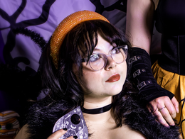 Portrait of comedian Mollia Risa wearing a ghost-patterned dress and holding a Ouija hand piece, with a halloween themed backdrop in the background