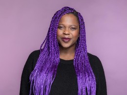 Studio portrait of comedian Tamara Shevon smiling toward the camera. Her vibrant braids match the colour of the wall behind her.