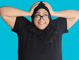 Portrait of comedian Monica Urribarri smiling with both her hands on her head, with a solid blue background.