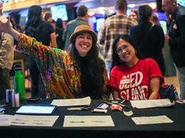 Two Sled volunteers sitting smile for the camera while sitting at a reception table for a Sled Island event in 2025.