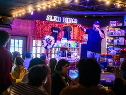 A burly host stands on stage and talks into the microphone at the Ship & Anchor during Punk Rock Bingo. A wall of prizes is behind him alongside a bright sign that reads "Sled Bingo."
