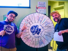 The two Quiz Shoe hosts smile for a photo in front of their prize wheel during a quiz event at Tubby's during Sled Island 2024.