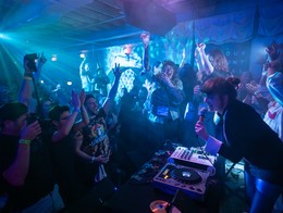 A colour photo shows Annie-Claude Deschanes playing an intimate show in front of a very enthusiastic crowd. They are seen singing into a microphone, behind a turntable. Everyone is bathed in blue light.