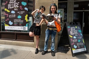 Photo of two festival attendees looking through the Sled Island Program Guide at the front door entrance of SpanicArts. A large decal and sandwich board with Sled Village information are displayed beside them.