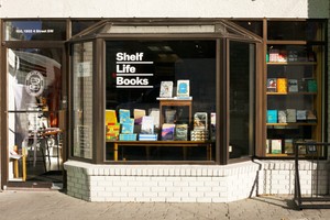 A colour photo shows a street view of the front door and bay window of Shelf Life Books. White decals with the business name are seen on the door and window, and you can see books on display through the glass.