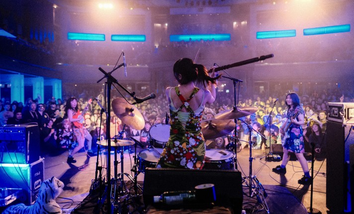 A dynamic live music performance from Otoboke Beaver captured from behind the drummer, who is wearing a colorful floral dress and energetically playing a drum kit on stage. To the left, a guitarist strums an electric guitar, while a bassist plays to the right. Blue lighting bathes the scene, highlighting the packed audience in the background, many of whom are cheering and raising their hands. The venue features tiered seating and a lively atmosphere, with musical gear and stage equipment surrounding the band.
