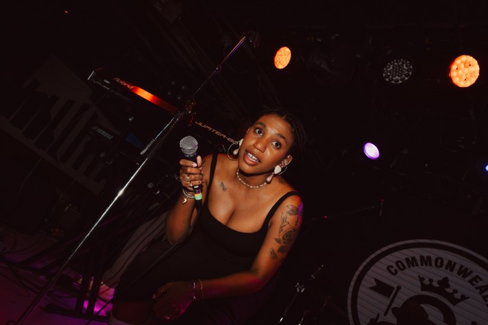Singer Yaya Bey stands on stage holding a microphone, dressed in a black sleeveless dress with layered bracelets and visible tattoos on the left arm. Behind them, a keyboard labeled "Jupiter" is positioned among stage equipment, illuminated by concert lighting. The background features a circular emblem with the word “COMMONWEALTH” and a crown symbol, which is the venue. The atmosphere is intimate and focused, capturing a powerful live performance moment.