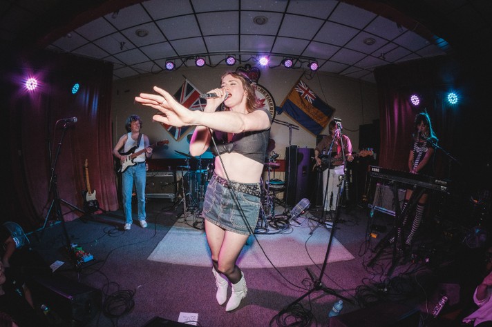 A lively concert scene with the five-member band TOPS performing on a brightly lit stage. The lead singer stands in front, wearing a black crop top, denim skirt, and white boots, singing into a microphone with one arm extended outward. Other band members—guitarist, bassist, drummer, and keyboardist—play their instruments under purple and blue spotlights. Colorful flags adorn the back of the stage, and cables and equipment are scattered across the floor, enhancing the raw, high-energy vibe of the performance.