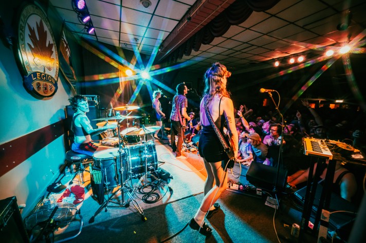 A high-energy concert scene with rock band Tropical Fuck Storm performing on stage. The drummer sits at a drum set on the left side, partially obscured by cymbals and lighting. In the foreground, a guitarist faces the crowd, while the bassist and another guitarist stand farther back. Bright beams of colorful stage lighting streak across the venue, creating a dramatic atmosphere. The crowd is engaged, cheering with raised arms. Behind the stage, a large wall emblem displays a maple leaf and the word “LEGION,” suggesting the venue's name or theme. Cables and music gear are scattered around the stage.