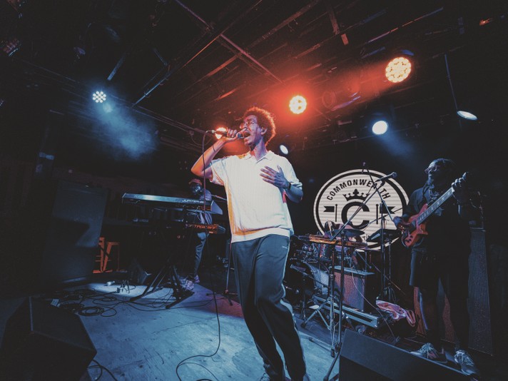 A live music performance from Oddisee & Good Compny on a dimly lit stage, awash in atmospheric blue and red lighting. The central performer holds a microphone and sings passionately. Behind them, a keyboardist and guitarist play amid scattered musical equipment. The background features a circular emblem displaying the word “COMMONWEALTH” and a stylized crown, suggesting the venue or event branding. The overall ambiance is moody and immersive, capturing the energy of a concert in full swing.