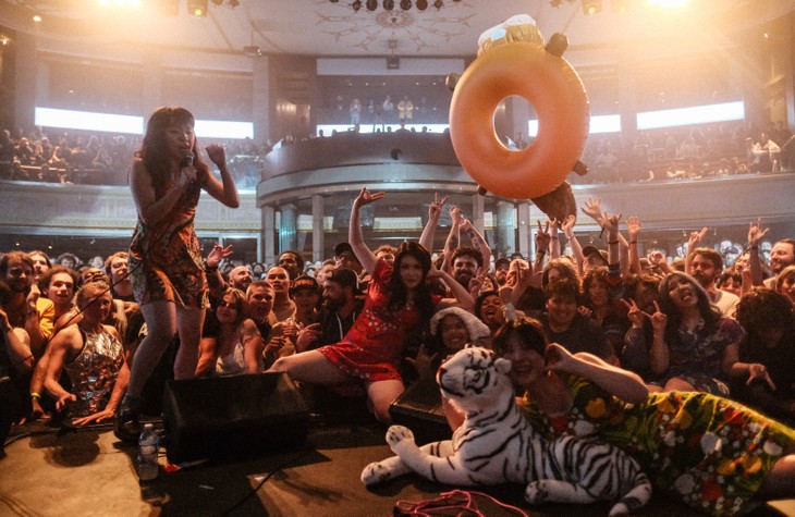 A vibrant concert scene with the band Otoboke Beaver, where the guitar player is seen on stage holding a microphone, surrounded by an enthusiastic crowd. Several people in the audience raise their hands, and an inflatable beaver donut floats above them. A large stuffed tiger lies on the edge of the stage in the foreground. The setting is festive and energetic, capturing the joyful atmosphere of a live music event.