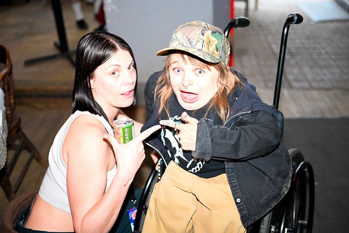 Two people pose together indoors, smiling and pointing at each other in a playful moment during Sled Island 2026. The person on the left has straight dark hair, wears a light gray tank top, and holds a can. The person on the right, seated in a wheelchair, wears a camouflage baseball cap, a dark jacket, and tan pants, with a surprised and expressive look on their face. The bright lighting highlights their joyful interaction and friendly energy.