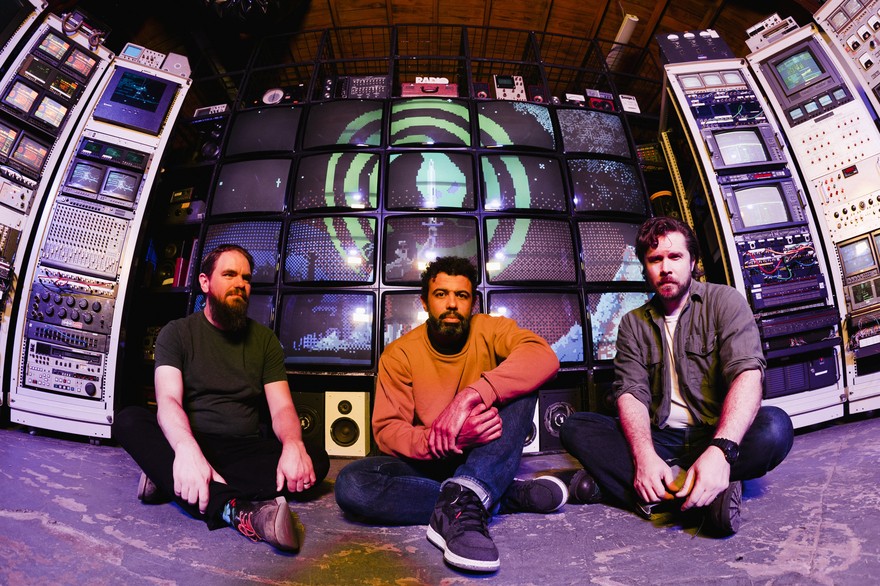 Photo of trio clipping., each sitting with legs crossed in front of multiple screens and audio-visual equipment.