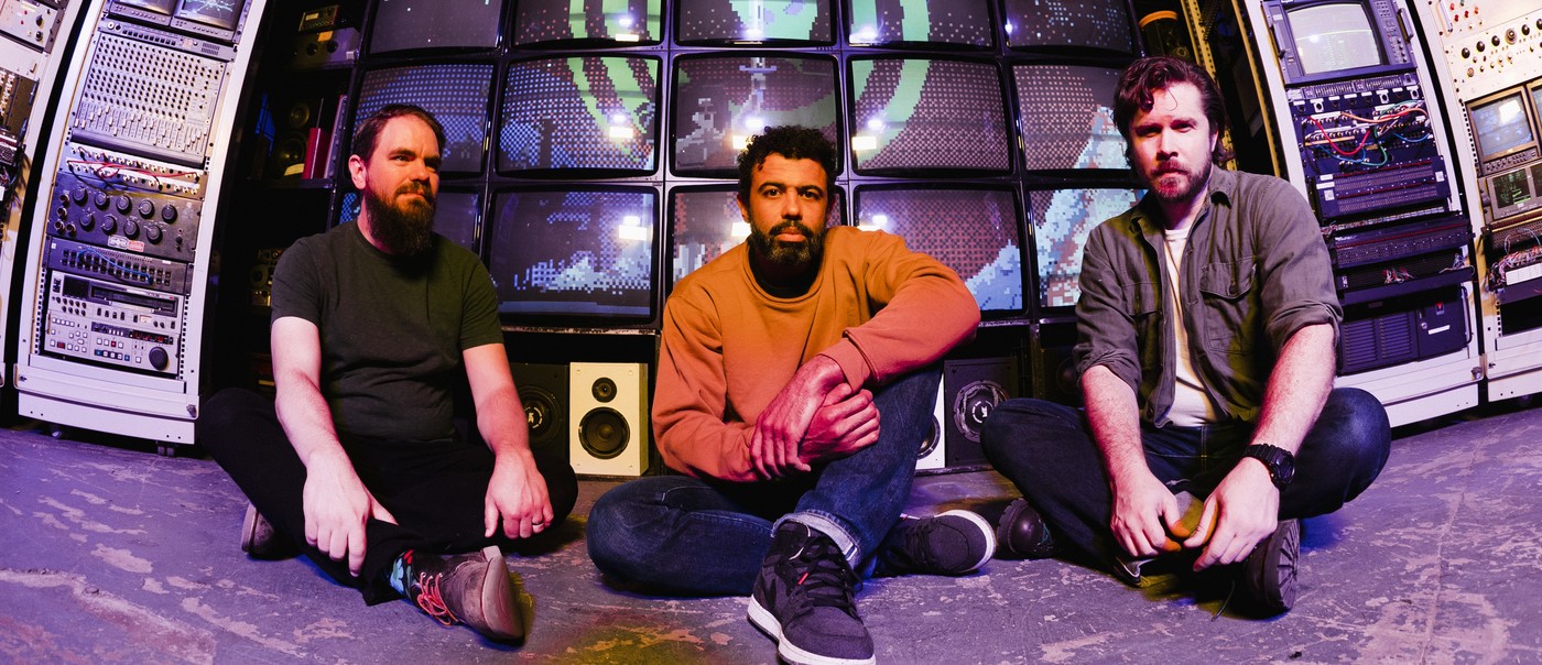 Photo of trio clipping., each sitting with legs crossed in front of multiple screens and audio-visual equipment.