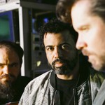 Three members of clipping. stand indoors in a dim, mixed‑tone studio space, surrounded by electronic equipment and monitors. The central figure looks directly at the camera while the others stand to the sides, one closer to the lens and slightly out of focus, creating an atmospheric, behind‑the‑scenes portrait.