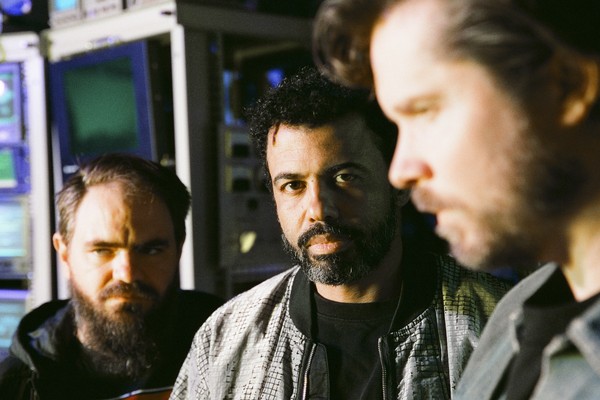 Three members of clipping. stand indoors in a dim, mixed‑tone studio space, surrounded by electronic equipment and monitors. The central figure looks directly at the camera while the others stand to the sides, one closer to the lens and slightly out of focus, creating an atmospheric, behind‑the‑scenes portrait.