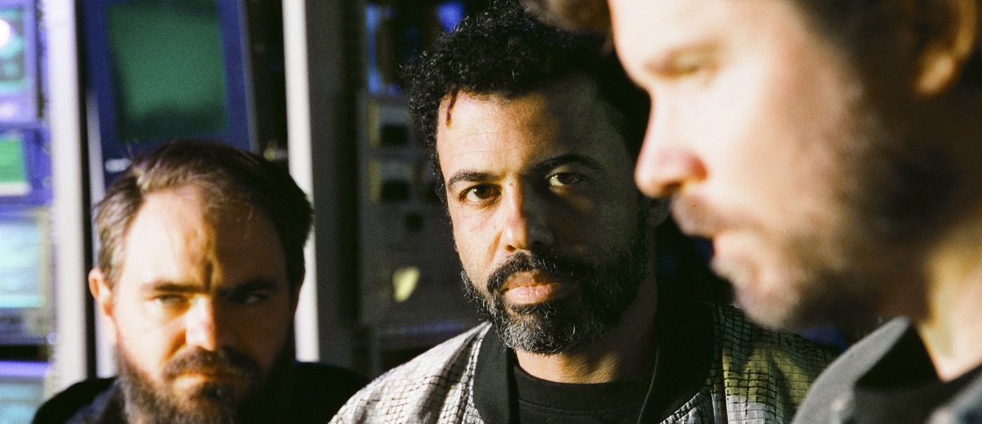 Photo of trio clipping., each sitting with legs crossed in front of multiple screens and audio-visual equipment.