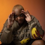 Rapper DijahSB is posed seated, with their hands on their head, with a warm backdrop behind them.